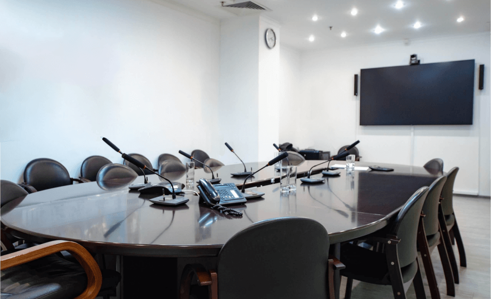 A conference room setting showcases the comparison of Huawei IdeaHub vs Traditional VC Systems. A large, oval wooden table is surrounded by multiple black chairs. Microphones are positioned on the table. A large screen, possibly a Huawei IdeaHub, is mounted on the wall.