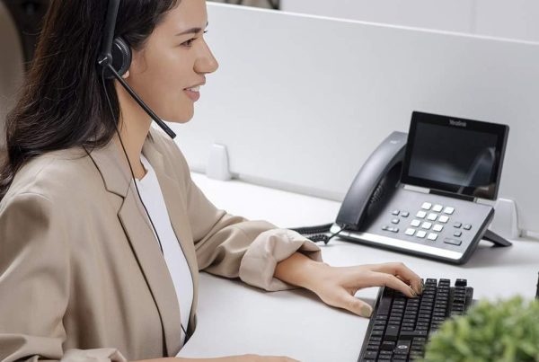 Smiling woman in a beige blazer using a wired headset while working on a computer at an office desk with a Yealink phone beside her.