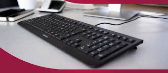 A black wired keyboard placed on a desk in an office setting, connected to a computer with a monitor and other accessories in the background.