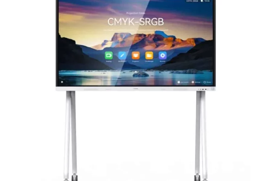 A front view of a large, white interactive display screen with a wide white bezel, mounted on a white mobile stand with four visible wheels. The screen displays a colorful landscape image with the text "CMYK-SRGB" centered on it. Below the text, there are several application icons, including a green "D" icon, a white brush icon, and a blue folder icon. A small camera is visible at the top center of the display's bezel.