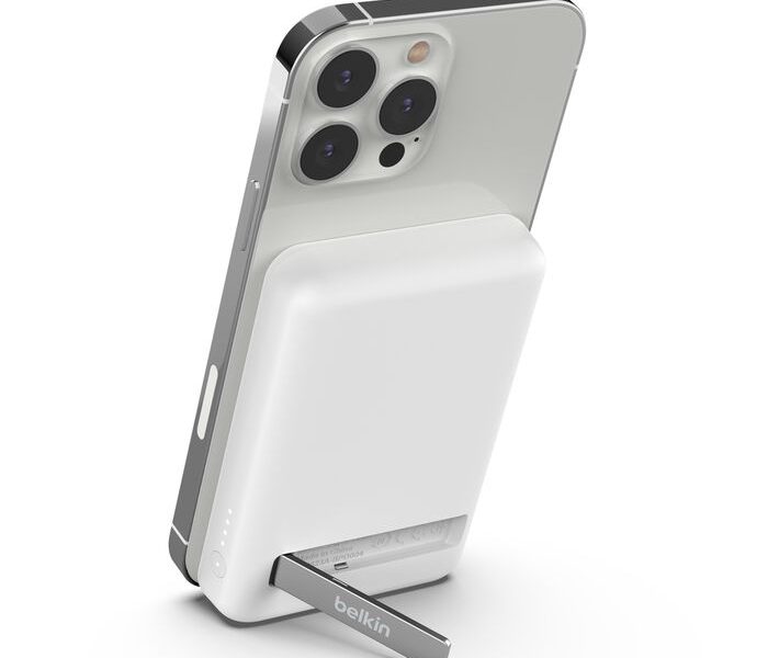 A white Belkin magnetic wireless power bank with a silver kickstand attached to the back of a silver iPhone. The power bank has rounded edges and an LED indicator with four lights visible on its bottom left side. The iPhone is slightly angled, showing its three camera lenses and the side button. The background is a solid white.