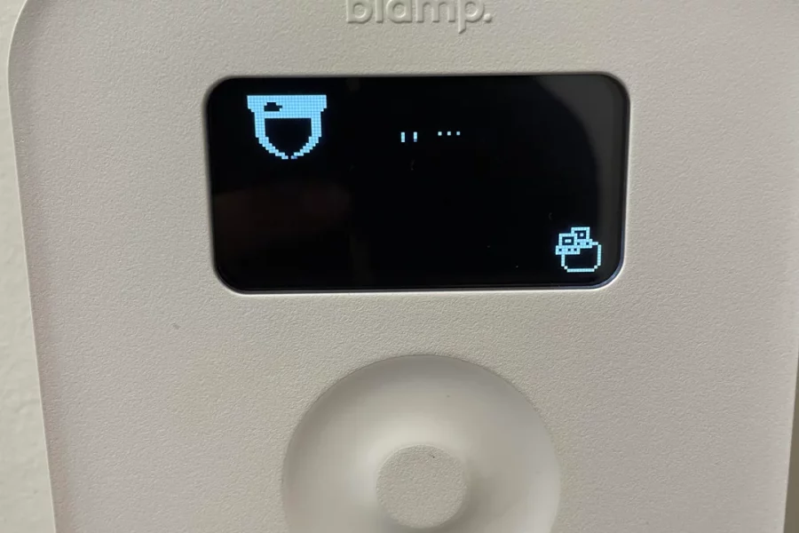 A close-up of a white Biamp Tesira TEC-1i control panel mounted on a wall. The black screen displays a pixelated bucket icon on the left, an ellipsis in the center, and a box icon with an arrow pointing out of it on the right. Below the screen is a circular, textured control dial.
