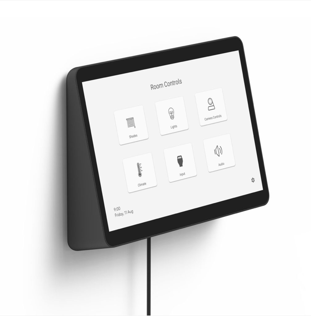 A black, wall-mounted smart room controller with a touch screen, angled on a light gray wall. The screen displays "Room Controls" and has icons for Shades, Lights, Camera Controls, Climate, Input, and Audio. A black cable extends from the bottom of the device.