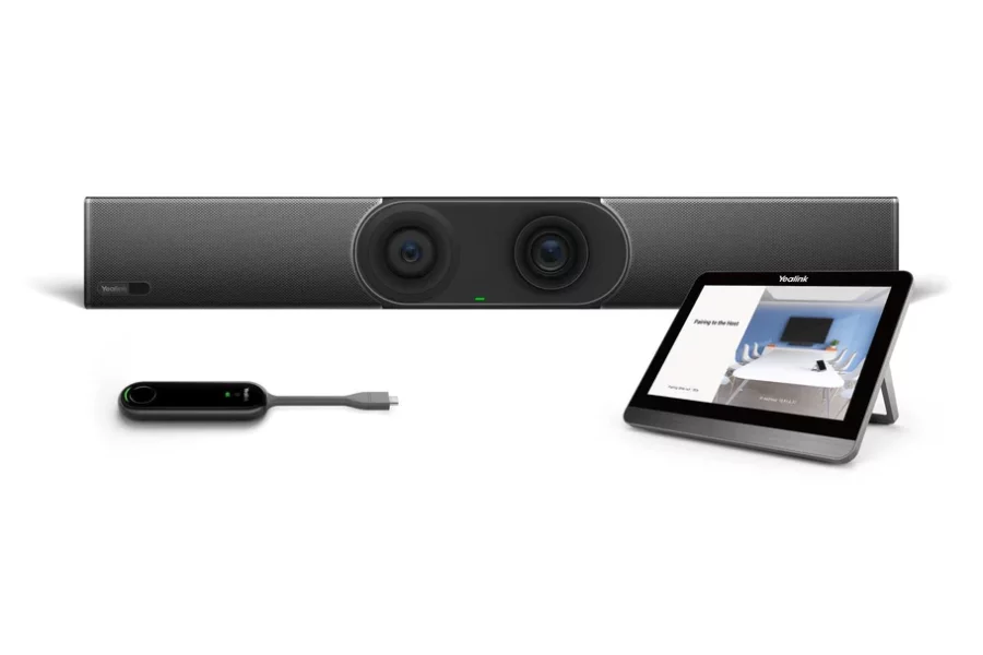 A video conferencing setup is presented against a white background. At the center, a long, horizontal video bar with two prominent camera lenses in the middle and speaker grilles on either side is positioned. The Yealink logo is visible on the far left of the video bar. To the right, a tablet with a black screen, displaying an image of a meeting room, is propped up on a stand. To the left, a sleek, black remote control with a glowing green indicator light and a short cable extends from its right side.