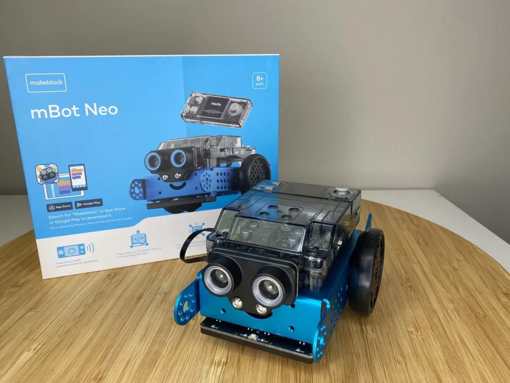 A blue makeblock mBot Neo robot with a clear top casing is positioned in the foreground on a light wooden surface. Its two large circular "eyes" with silver rims are prominent, and a subtle smile is designed below them. Behind the robot, its light blue and white retail box stands upright, displaying an image of the robot, the "makeblock" logo, and "mBot Neo" text, along with an "8+ AGES" label and icons for App Store and Google Play downloads.
