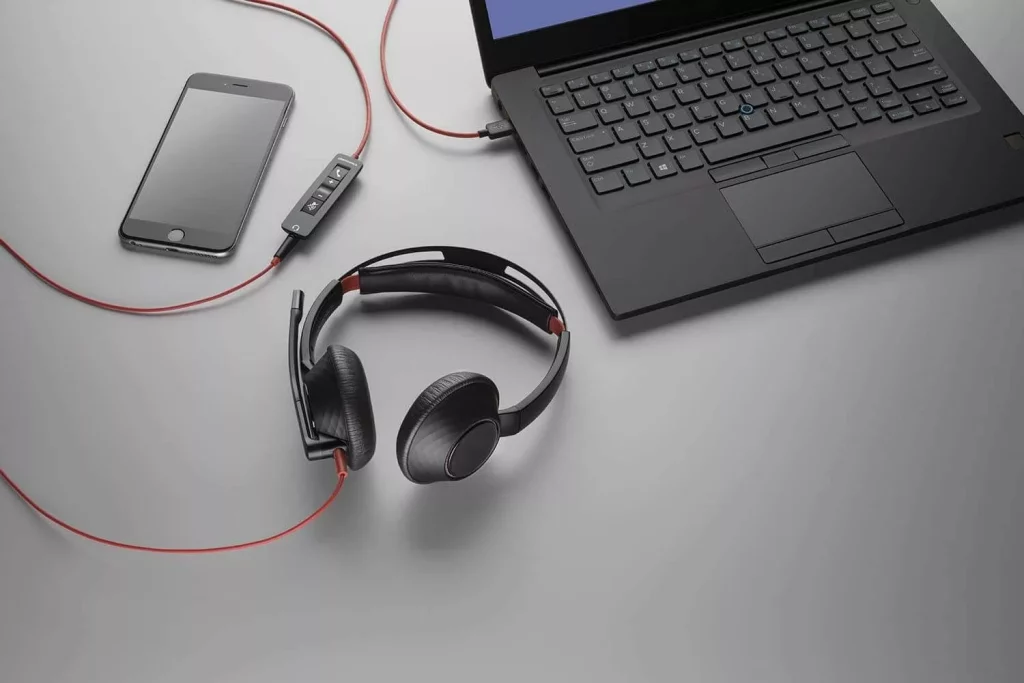 A flat lay, high-angle shot captures a black laptop, a smartphone, and a black headset with red accents on a gray surface. The laptop is positioned in the upper right, partially cropped, with a black cable extending from its right side. A black smartphone with a dark screen lies in the upper left. A black and red headset with an attached microphone boom is in the center foreground, its red cable snaking towards a small black inline control panel with buttons, which then connects to the laptop via a red cable. Another red cable from the control panel extends towards the smartphone.
