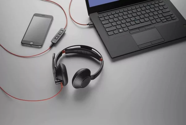 A flat lay, high-angle shot captures a black laptop, a smartphone, and a black headset with red accents on a gray surface. The laptop is positioned in the upper right, partially cropped, with a black cable extending from its right side. A black smartphone with a dark screen lies in the upper left. A black and red headset with an attached microphone boom is in the center foreground, its red cable snaking towards a small black inline control panel with buttons, which then connects to the laptop via a red cable. Another red cable from the control panel extends towards the smartphone.
