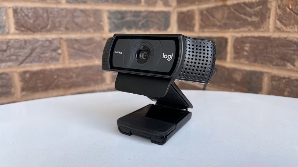 Black Logitech C920 webcam resting on a white surface with a brick wall background, angled slightly to the right with its mounting clip unfolded as a desktop stand.