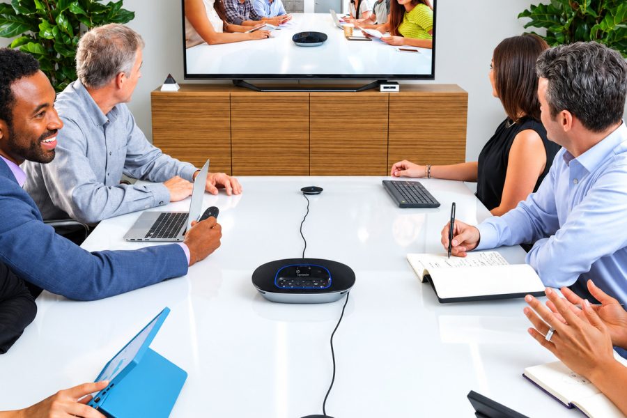 A group of people sitting around a conference table using a Logitech Group video conferencing system with a central speakerphone, expansion mics, and a large display screen showing remote participants.