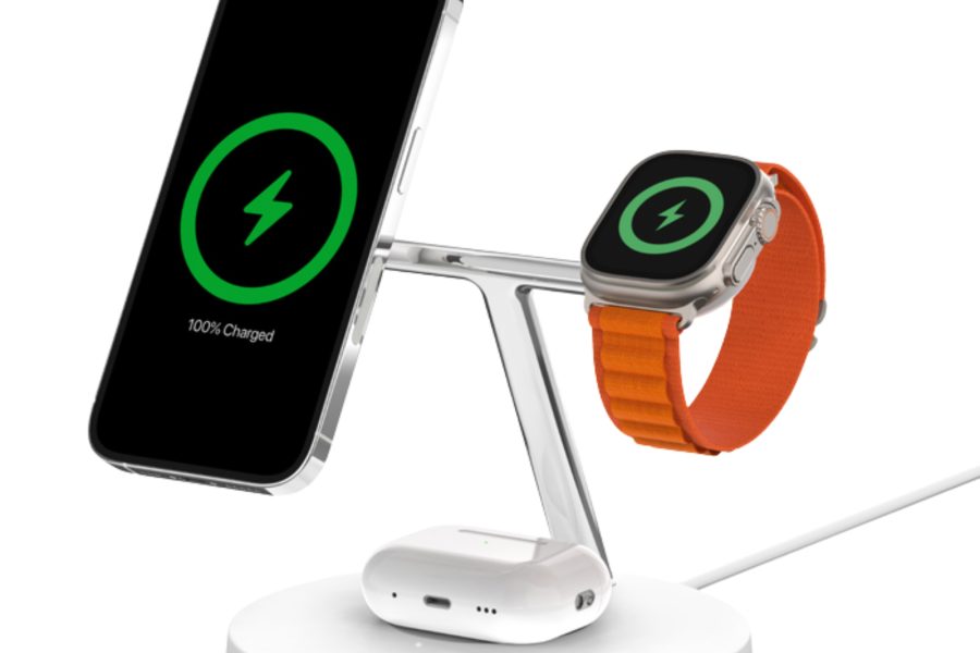 Belkin BoostCharge Pro 3-in-1 Wireless Charging Stand with MagSafe featuring an iPhone, Apple Watch Ultra with an orange strap, and AirPods charging simultaneously on a white base.