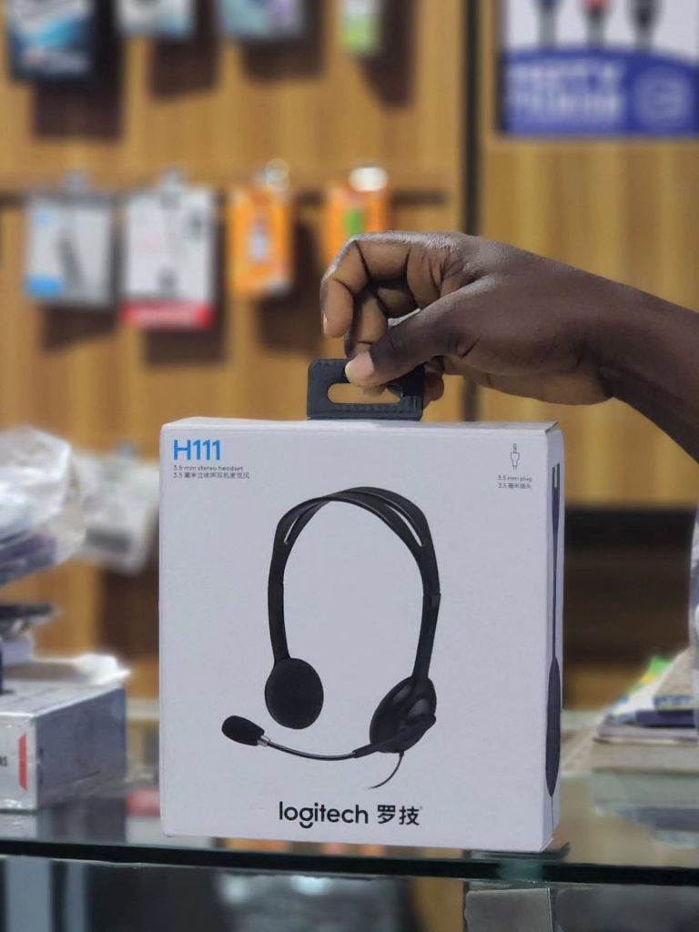 A hand holding the retail packaging box of a Logitech H111 3.5mm stereo headset in a store setting.