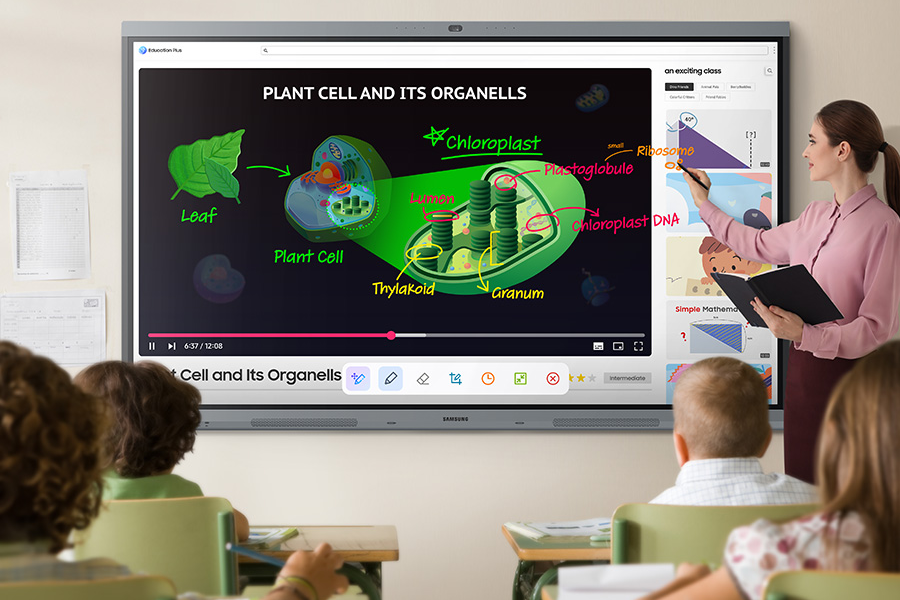 A teacher stands beside a large Samsung interactive display in a classroom, using a stylus to write on an educational lesson about plant cells and organelles while young students watch from their desks.