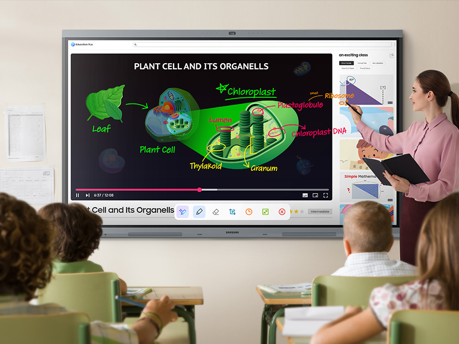 A teacher stands beside a large Samsung interactive display in a classroom, using a stylus to write on an educational lesson about plant cells and organelles while young students watch from their desks.